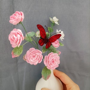 May include: A white ceramic vase with a round base holds a bouquet of crocheted pink roses and a red crocheted butterfly.