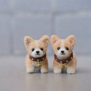 May include: Two small, brown and white felt corgi dog figurines with brown collars and gold bells. The corgis are standing on a gray surface.