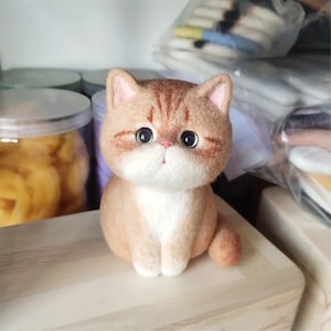 May include: A felted orange and white cat figurine with a white chest and a white face. The cat has black eyes and a pink nose.