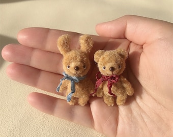 May include: Two small plush toys, a brown bunny and a brown bear, are sitting in the palm of a hand. The bunny has a blue ribbon around its neck and the bear has a red ribbon around its neck.