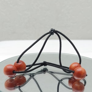 May include: A unique hair accessory featuring six reddish-brown wooden beads strung on black elastic cords. The beads are arranged in a geometric pattern, creating a distinctive and eye-catching design. The accessory is displayed on a reflective surface.