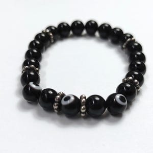 May include: A black beaded bracelet featuring a combination of solid black and eye-shaped beads. The eye beads have a white iris and a black pupil. Silver-coloured spacers are between some of the beads. The bracelet is on a white background.