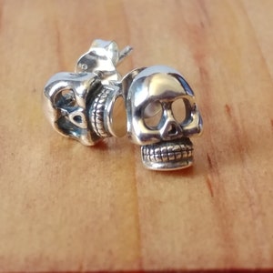 May include: A pair of polished silver stud earrings shaped like human skulls. Each earring features detailed eye sockets, a nasal cavity, and a textured jawline. The silver metal has a reflective finish, and the posts are visible on the back of the skulls.