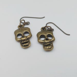 May include: A pair of antique brass-colored skull earrings. Each earring features a detailed skull pendant with open eye sockets, a nose cavity, and a full set of teeth. The earrings have a simple hook design.