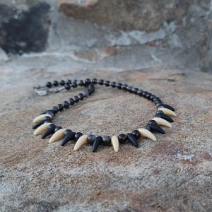 May include: A necklace featuring a series of black and off-white pointed elements, alternating with small black beads. The necklace is displayed on a textured, light brown stone surface.