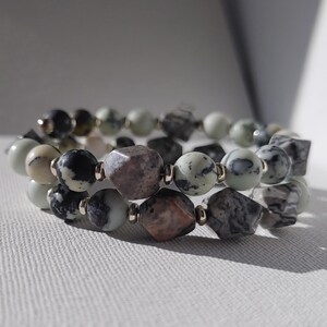 Könnte beinhalten: Ein mehrreihiges Wickelarmband mit einer Vielzahl von runden und facettierten Perlen in Schwarz-, Weiß-, Grau- und gedeckten Rosatönen. Die Perlen sind mit kleinen metallischen Zwischenelementen durchsetzt, was für eine strukturierte Optik sorgt.