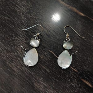 May include: A pair of silver cat earrings. Each earring features a cat head and a pear-shaped body, both made of a pearlescent material. The earrings have a curved tail and a silver hook.