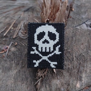May include: A black and white beaded patch featuring a skull and crossbones design. The skull is white, and the background is black. The patch is rectangular and likely handmade, with a detailed beadwork pattern.