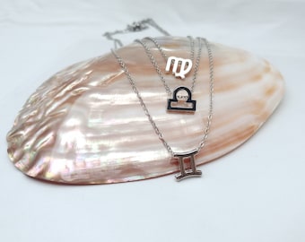 May include: Three silver necklaces with zodiac signs: Virgo, Libra, and Gemini. The necklaces are displayed on a white mother-of-pearl shell.