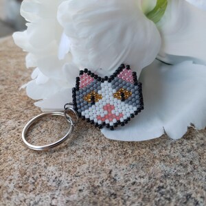 May include: A beaded cat keychain with a silver keyring. The cat's face is white, gray, pink, and black, with gold-colored eyes. The keychain is set against a white flower and a textured, tan surface.