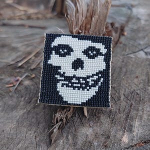 May include: A square, beaded patch featuring a white skull design on a black background. The skull has large eyes and a wide grin. The patch is likely an accessory, possibly for clothing or bags.