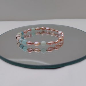 May include: A delicate bracelet featuring a combination of pink and light blue beads. The bracelet is made with freshwater pearls and aquamarine beads, strung together with small metal accents. The bracelet is displayed on a reflective surface.