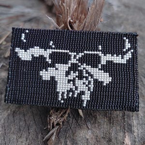 May include: A rectangular, beaded wallet in black with a white scorpion design. The scorpion is centered on the wallet and is made of small, white beads. The wallet is likely handmade and has a unique, artistic appearance.