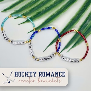 May include: Three beaded bracelets in blue, red, and teal, with gold accents. Each bracelet features white letter beads spelling out names and phrases. The image includes text that reads "Hockey Romance reader bracelets."