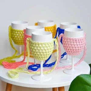 Könnte beinhalten: Eine Sammlung weißer Weingläser aus Kunststoff, die jeweils in einer farbenfrohen, gehäkelten Netzhülle mit langem Trageriemen stecken. Die Hüllen sind in leuchtendem Limettengrün, Pink, Gelb und Königsblau gehalten und haben ein Gittermuster.