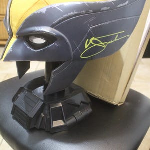 Peut inclure: Un casque de personnage Wolverine avec une partie supérieure jaune et un masque facial gris foncé doté d'ailerons latéraux pointus. Le masque présente une signature jaune vif sur le côté gauche et est monté sur un support noir à base géométrique.
