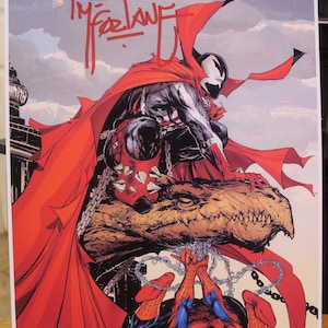 May include: A comic book art print featuring Spawn in a black and white suit with a long red cape, crouching on a stone gargoyle, while Spider-Man hangs upside down below, entangled in silver chains. The print includes a red Todd McFarlane signature.
