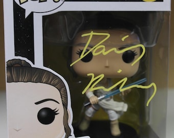 May include: A white and yellow Star Wars Pop! figure of Rey from the movie Star Wars: The Force Awakens. The figure is holding a blue lightsaber. The box is white with black and yellow lettering. The number 307 is printed on the box. The figure is signed by Daisy Ridley.