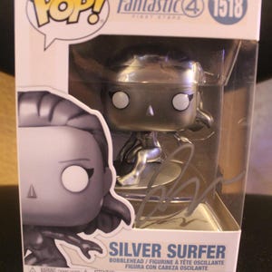 Puede incluir: Figura de Silver Surfer Funko Pop! en una vitrina de plástico transparente. La figura es plateada con ojos blancos y está sobre una tabla de surf. La caja tiene el logo de los Cuatro Fantásticos y el número 1518.