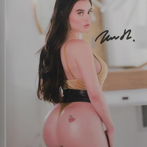 May include: A glossy photograph featuring a person with long dark hair wearing a gold-colored bodysuit and a black belt. The person is looking over their shoulder, and a small tattoo of two red hearts is visible. A black ink signature is on the image.