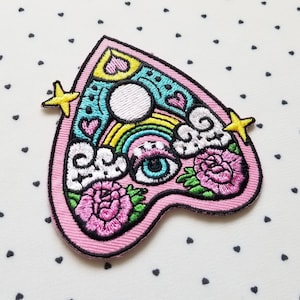 May include: A pink, heart-shaped embroidered patch with a whimsical design. The patch features a white moon, rainbow, eye, flowers, and stars. The design is outlined in black and has a pink border. The background is white with small black hearts.