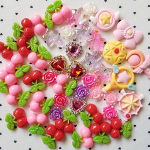 May include: A collection of colorful resin embellishments, including cherries, roses, stars, hearts, and other decorative shapes. The embellishments are in shades of red, pink, purple, and yellow, with some featuring glitter and rhinestones. These are ideal for crafting and scrapbooking.