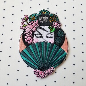 May include: Embroidered patch featuring a geisha with closed eyes, a fan, and floral accents. The geisha's hair is black, and the fan is teal. Pink flowers and a peach-colored oval frame the geisha.