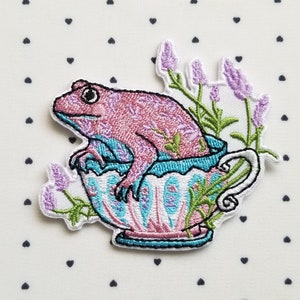 May include: Embroidered patch of a pink frog sitting in a teacup with lavender flowers. The teacup is decorated with a blue and pink floral pattern.
