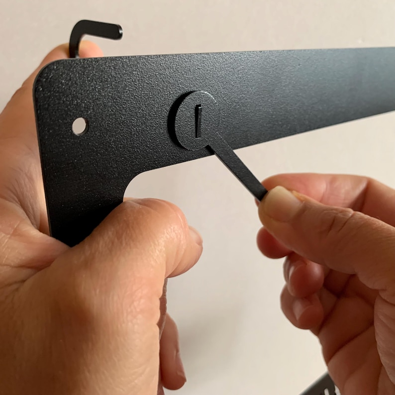 Pu&ograve; includere: Un supporto da parete in metallo nero con un piccolo foro rotondo e un gancio in metallo. Il gancio viene sollevato da un dito.
