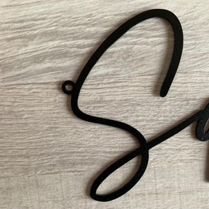 May include: Black metal cutout of the letter "S" in a cursive font. The letter is attached to a wall with a small hole at the top.