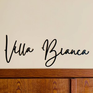 May include: Black metal wall decor with the words "Villa Bianca" in a cursive script font.