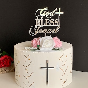 Op de afbeelding: Witte cake met gouden accenten versierd met een zilveren caketopper waarop "God Bless Jonacl" staat met een kruis. De cake is ook versierd met roze en witte bloemen.