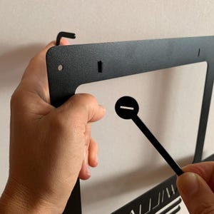 Pu&ograve; includere: Telaio in metallo nero con un gancio in alto e un piccolo pezzo di plastica nera con una fessura. Il telaio &egrave; tenuto da una mano.