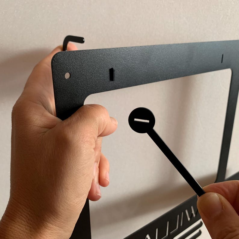 Pu&ograve; includere: Telaio in metallo nero con un gancio in alto e un piccolo pezzo di plastica nera con una fessura. Il telaio &egrave; tenuto da una mano.
