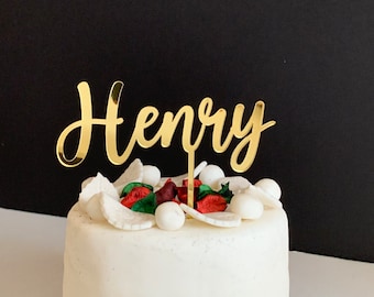 Può includere: Topper per torta dorato con il nome "Henry" scritto in corsivo. Il topper è posto sopra una torta bianca decorata con glassa bianca e rossa.