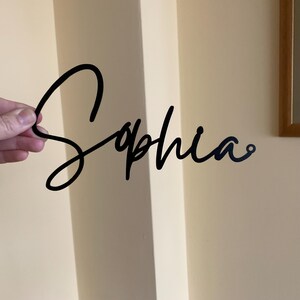 May include: Black metal cutout of the name "Sophia" in a cursive script font.