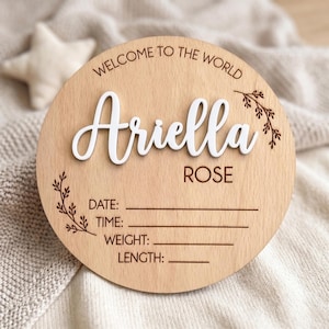 Könnte beinhalten: Ein rundes Geburtsankündigungsschild aus Holz mit dem lasergravierten Text WELCOME TO THE WORLD und dem Namen Ariella ROSE in erhabenen weißen Buchstaben. Es enthält gravierte florale Akzente sowie Linien für Datum, Uhrzeit, Gewicht und Länge.