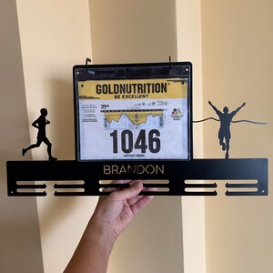 Pu&ograve; includere: Porta medaglie in metallo nero con una silhouette di corridore su ciascuna estremit&agrave;. Il supporto ha un porta pettorale in plastica trasparente con il testo "Goldnutrition Be Excellent" e il numero "1046". Il nome "Brandon" &egrave; stampato sul supporto.