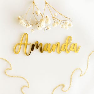 Op de afbeelding: Een goudkleurig naamplaatje in cursieve stijl met de naam "Amanda" staat centraal in deze afbeelding. Het naamplaatje is geplaatst tegen een witte achtergrond, aangevuld met delicate witte bloemen en goudkleurige touwaccenten. De algehele esthetiek is elegant en minimalistisch.