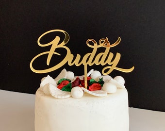 Può includere: Topper per torta dorata con la parola "Buddy" in corsivo. Il topper è posto sopra una torta bianca decorata con pasta di zucchero bianca e rossa.