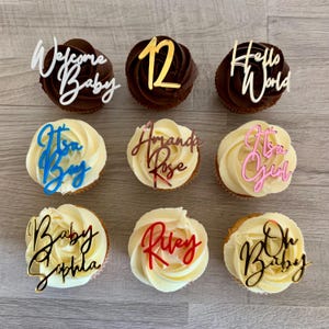 Può includere: Nove cupcake decorati con topper in acrilico dorati, rosa, rossi e blu. I topper hanno varie frasi, tra cui "Welcome Baby", "It's a Boy", "It's a Girl", "Oh Baby", "Riley", "Baby Sophia", "Amanda Rose", "Hello World" e "12".