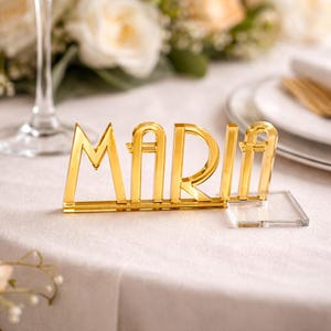 Könnte beinhalten: Eine goldfarbene Tischkarte mit dem Namen "MARIA" in einem auffälligen Art-Deco-Stil. Die Buchstaben sind an einer transparenten, rechteckigen Basis befestigt. Der Hintergrund zeigt eine weiße Tischdecke und Blumengestecke, passend für eine Hochzeit.