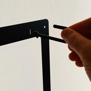 Pu&ograve; includere: Un telaio in metallo nero con un piccolo pezzo di plastica nero attaccato ad esso. Una mano tiene uno strumento in metallo nero e lo sta usando per stringere il pezzo di plastica.