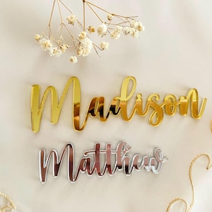 Może przedstawiać: Złote i srebrne wycięcia imion, "Madison" i "Matthew", w kursywie. Imiona są wyświetlane na białej powierzchni, z suszonymi kwiatami i złotymi akcentami sznurkowymi. Idealne do dekoracji ślubnych lub spersonalizowanych prezent&oacute;w.