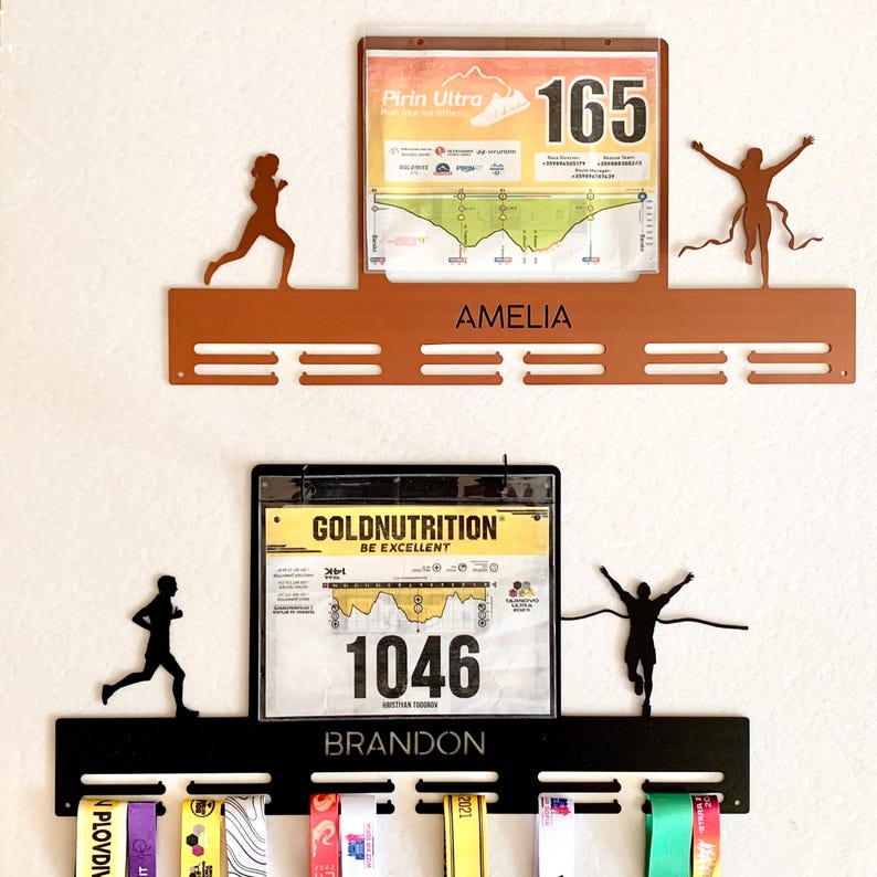 Pu&ograve; includere: Due porta medaglie in metallo nero con silhouette di corridori. Il supporto superiore &egrave; marrone e ha un pettorale da corsa con il testo "Pirin Ultra" e il numero "165". Il supporto inferiore ha un pettorale da corsa con il testo "Goldnutrition Be Excellent" e il numero "1046". Il supporto superiore &egrave; etichettato "Amelia" e il supporto inferiore "Brandon".