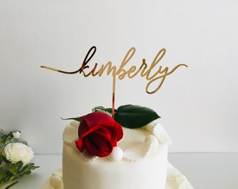 Può includere: Topper per torta dorato con il nome "Kimberly" scritto in corsivo. Il topper è posizionato su una torta bianca decorata con una singola rosa rossa e glassa bianca.