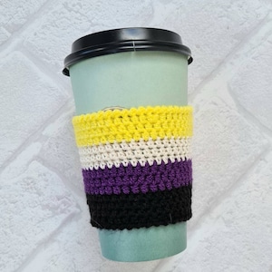 Puede incluir: Un calentador de taza de café de ganchillo con rayas amarillas, blancas, moradas y negras. El calentador está en una taza para llevar verde.