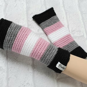 May include: A pair of crocheted fingerless gloves in black, grey, white and pink stripes. The gloves are on a white brick background. The gloves have a tag that says "Pride".