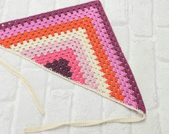 May include: A triangular crocheted scarf with a multi-colored gradient pattern in shades of pink, orange, and purple. The scarf has a white crocheted fringe around the edges and a long white crocheted tie.