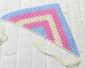 May include: A crocheted triangular scarf with a light blue, pink, and cream color pattern. The scarf has a cream-colored tie.